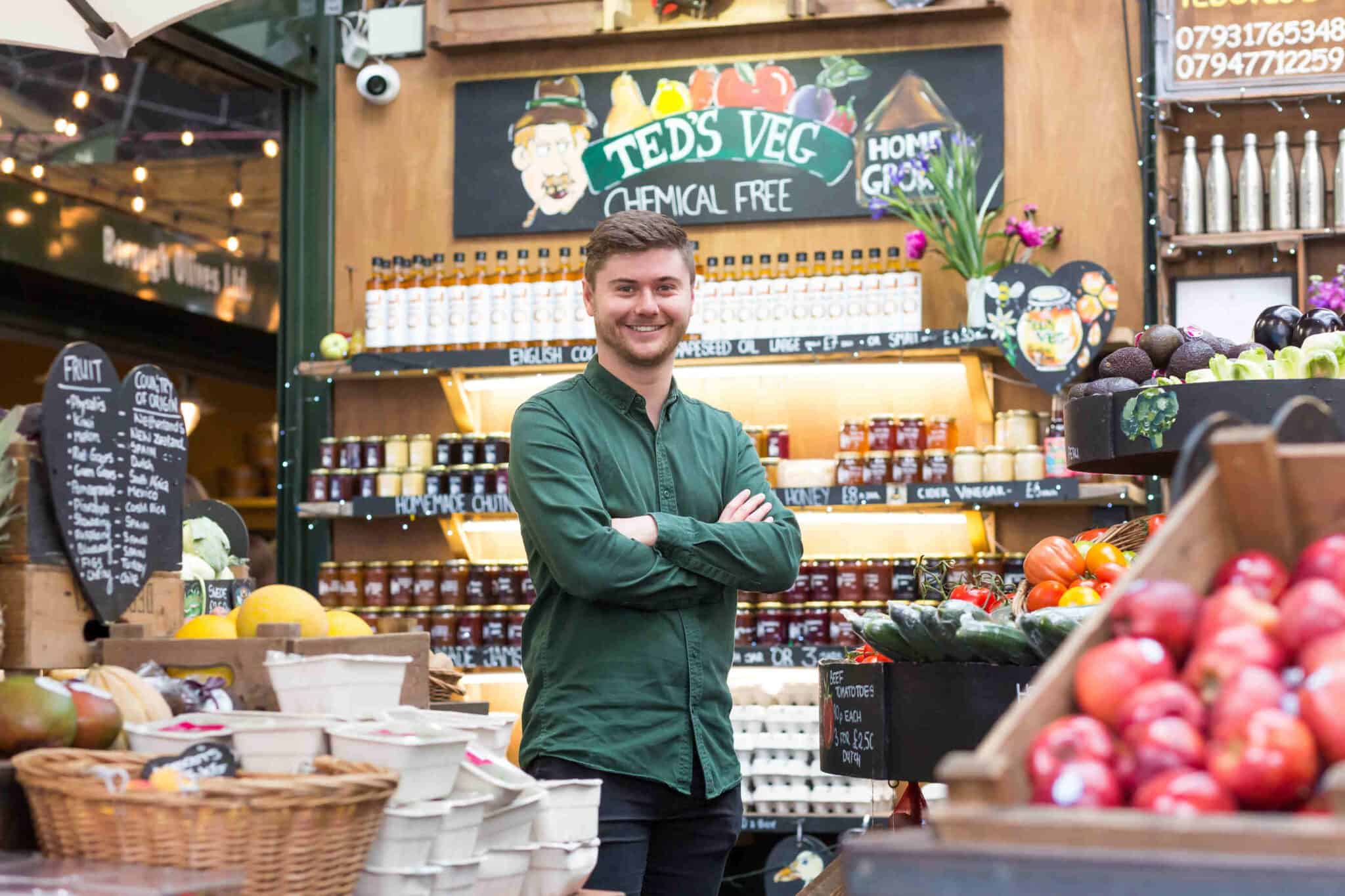 Sam is standing with his arms crossed in front of an array of fruit and vegetables. Behind him are 3 shelves laden with condiments and oils. He's wearing a green shirt and smiling.