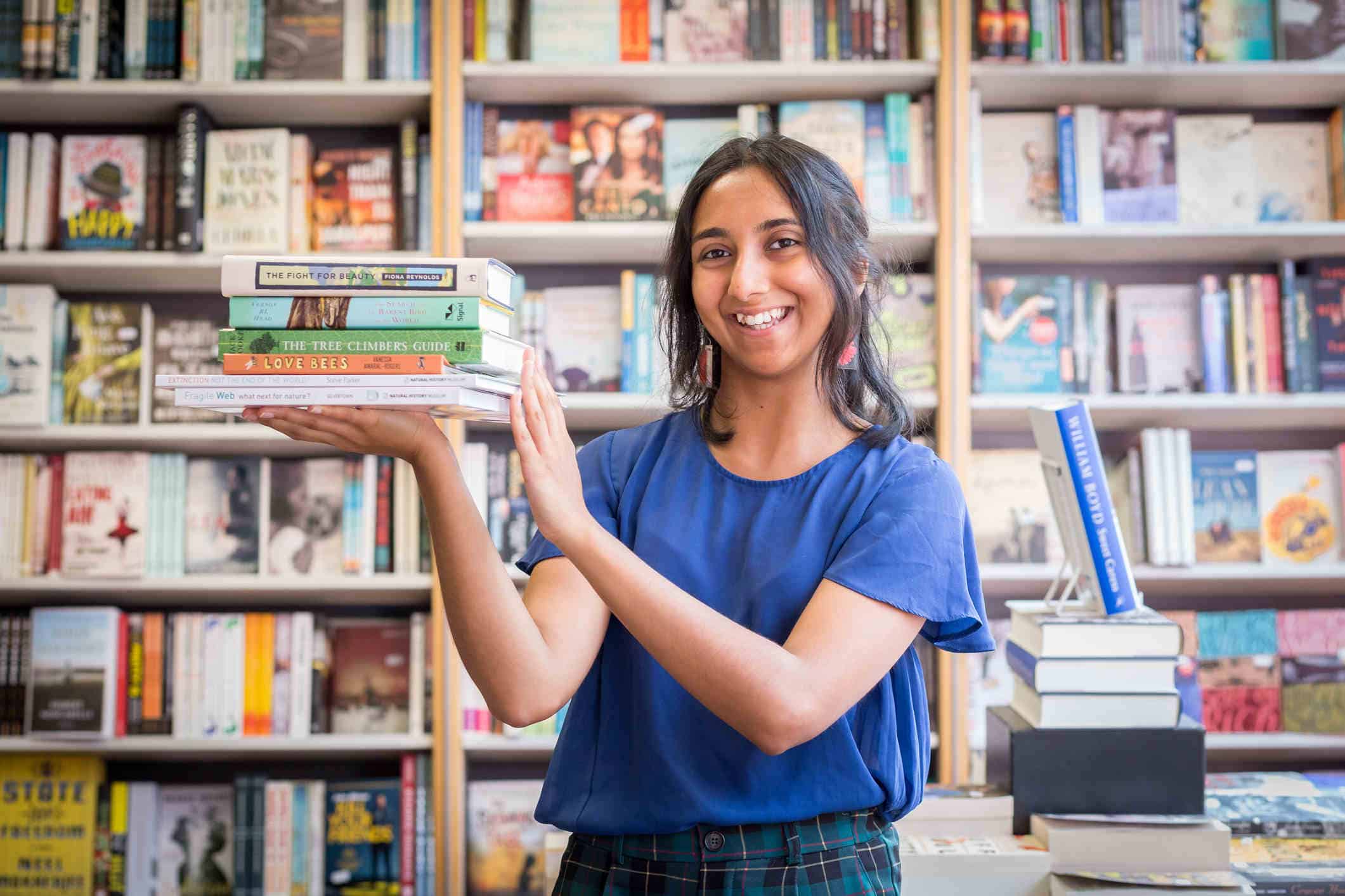 A brown person stands in a bookshop wearing a dark blue blouse. She is holding 5 books in her hand.
